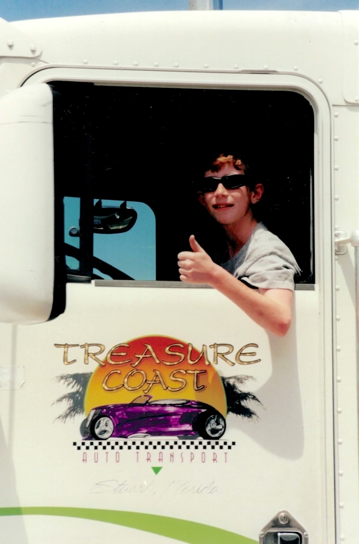 One of the Convente twins sitting in his Dad's 18 wheeler truck cab. The door window is down, and the twin is giving a thumb's up gesture. The door graphics read Treasure Coast Auto Transport, Stuart, Florida and have art of a purple hot rod car in front of a sunset.